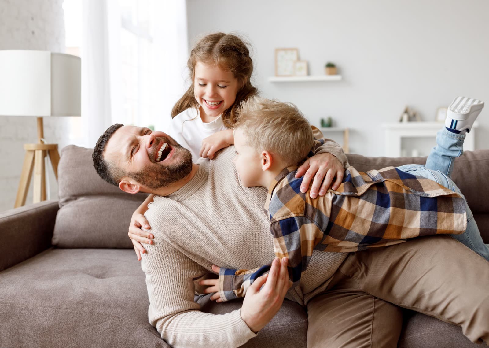 A father laughing on a sofa as his son and daughter climb over him